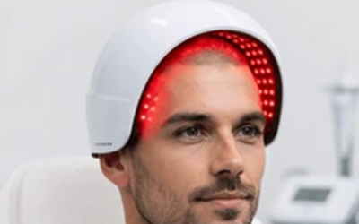 Man wearing a white LED light therapy helmet emitting red lights in a clinical setting.