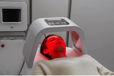 Person reclining inside a curved red LED light therapy device with head on a pillow in a treatment room.