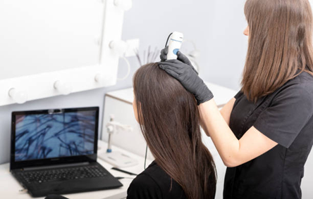 Hair stylist wearing black gloves uses a handheld scanner on a client's scalp while a laptop shows magnified scalp images.