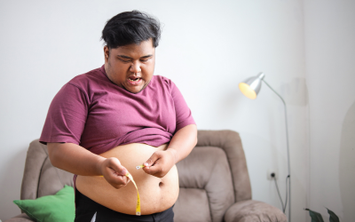 Man sitting on a sofa measures his waist with a yellow tape measure, wearing a maroon shirt and showing a focused expression.