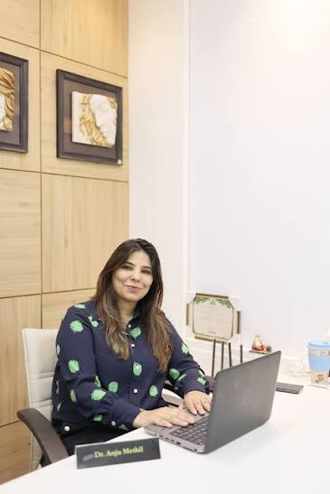 Dr. Anjo Merali sits at a white desk with a laptop in a modern clinic, smiling at the camera.