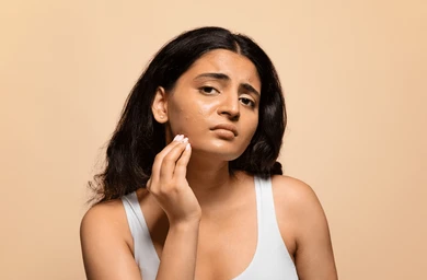 Woman with dark hair in a white tank top touches her cheek in a warm beige studio portrait.