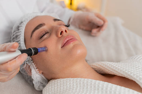 Picture2 Woman receiving a facial treatment with a handheld device on her cheek while eyes are closed, wearing a spa cap and robe in a calm setting.