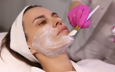 â_¢ Step 4_ Topical anaesthesia Woman receiving a facial; clinician in pink gloves brushes a white facial mask onto her face.