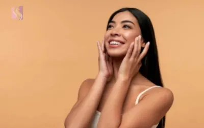 Smiling woman with long dark hair, touching her cheeks with both hands, wearing a white top against a peach background.