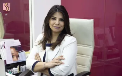 Professional woman in a white blazer seated at a desk in a modern office, smiling at the camera.