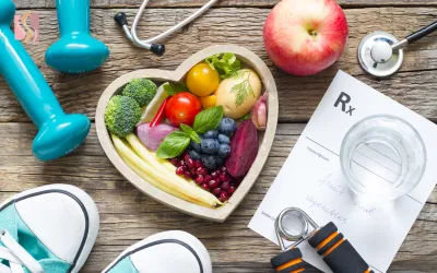 Heart-shaped bowl of colorful fruits and vegetables on a wooden table, surrounded by a stethoscope, a prescription form, water glass, and gym gear for healthy living.