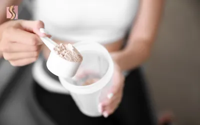 Person scooping protein powder into a white shaker cup held by a person in a white top